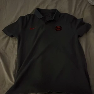 Svart PSG x Nike Dri-Fit fotbollströja - Svart PSG fotbollströja från Nike med röd Nike-logga och PSG-emblem på bröstet. Slim fit-modell i Dri-Fit-material som andas, perfekt för träning eller match. Klassisk pikékrage och korta ärmar ger en clean och sportig look.