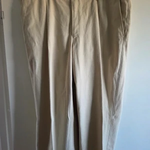 Beiga kostymbyxor i bomull C56 - Snygga beiga kostymbyxor i 100% bomull. Klassisk rak passform med pressveck och sidfickor. Perfekta för dig som gillar stilrena och enkla byxor med en clean look. Storlek C56 enligt lappen.