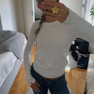 Vit långärmad tröja från Zara - Snygg vit långärmad tröja från Zara med rund halsringning och dekorativa guldfärgade knappar vid ärmsluten. Tröjan är i mjuk bomullsmix och har en slim passform som sitter snyggt till jeans!