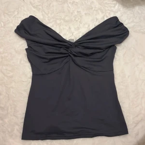 Svart offshoulder topp med twist - Snygg svart topp med offshoulder-design och twistad detalj framtill. Figurnära passform och mjukt, stretchigt material som sitter skönt. Perfekt för dig som vill ha en stilren men ändå unik look.