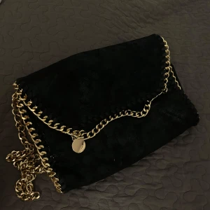 Svart kuvertväska med guldkedja - Stilren svart kuvertväska i mocka med snygga guldfärgade kedjedetaljer runt kanterna och som axelrem. Perfekt för att lyfta vilken outfit som helst med en lyxig touch. Väskan har ett lock med kedja och rund guldfärgad berlock.