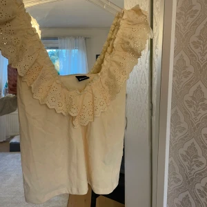 Gul blus med volang från Gina Tricot - Supersöt gul blus från Gina Tricot med bred volangkrage i broderad spets. Ärmlös modell med djup v-ringning och romantiska detaljer. Perfekt för dig som gillar en feminin och trendig look.