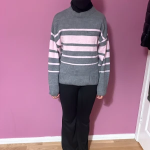 Grå och rosa randig stickad tröja - Säljer en mysig stickad tröja i grått med breda och smala rosa ränder. Tröjan har en rund hals, långärmade ärmar och ribbade muddar vid ärmslut och nederkant. Perfekt för dig som gillar chill och stilren look.