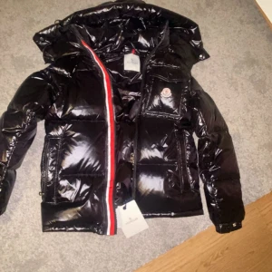 Svart glansig dunjacka från Moncler - Säljer en svart glansig dunjacka från Moncler med huva och klassisk logga på bröstfickan. Jackan har röda och vita detaljer längs dragkedjan, flera fickor med dragkedja och tryckknapp samt quiltad design. Perfekt för kalla dagar och riktigt snygg streetstil. Har inte använt den helt ny .