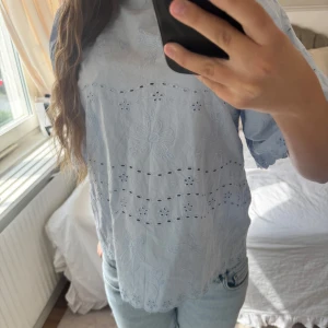 Ljusblå broderad blus med snörning - Supersöt ljusblå blus med broderade blommor och hålmönster. Blusen har korta ärmar, vacker öppen rygg med snörning och volangkant. Från Vero Moda.