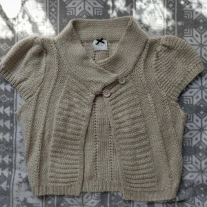 Beige stickad väst med kort ärm - Säljer en beige stickad väst från Lindex med korta puffärmar och bred krage. Västen har två knappar framtill och ribbade detaljer längs kanterna. Perfekt att slänga över en t-shirt för en chill look.