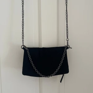 Svart axelväska med kedja - Snygg svart axelväska i mjukt tyg med coola metallkedjor som axelrem och handtag. Väskan har dragkedja upptill och en enkel, stilren design som passar till det mesta. Perfekt storlek för mobil, plånbok och nycklar. Väskan är från mango kids och köpt på Zalando .Öppen för prisförslag