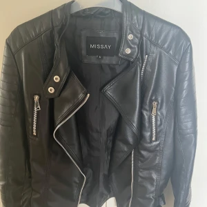 Svart skinnjacka från Missay, S - Snygg svart skinnjacka från Missay i bikerstil med silvriga dragkedjor och detaljer. Jackan har markerade axlar, flera fickor med dragkedja och coola sömmar på ärmarna. Perfekt för dig som vill ha en edgy look till vardags. Använd 2-3 gånger.