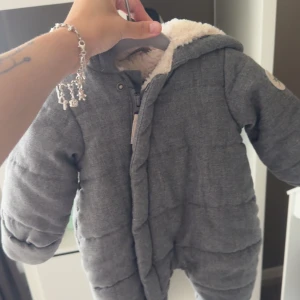 Grå pufferjacka med öron från C mini - Supermysig grå overall för baby från C mini, storlek 50/56. Jackan har fluffigt foder, huva med söta öron och dragkedja framtill. Perfekt för kalla dagar när du vill att din lilla ska vara både varm och gullig.