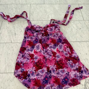 Blommig kortklänning med knytband från Adoore - Säljer en färgstark kortklänning med blommigt mönster i rosa, lila och rött från Adoore. Klänningen har knytband vid axlarna och en lätt A-linjeformad siluett. Perfekt för dig som gillar att sticka ut med mycket färg och mönster. Materialet känns lätt och luftigt.