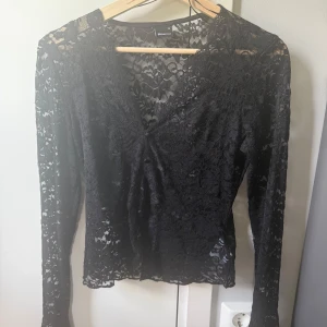 Svart spetsblus från Gina Tricot - Svart blus i genombruten spets från Gina Tricot med v-ringad omlottfront och dekorativa små knappar. Långärmad och figurnära passform, perfekt för dig som gillar detaljer och en lite mer dressad vibe. Spetsen har ett blommigt mönster som ger en extra touch.