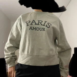 Grön sweatshirt med pärlor - Grön sweatshirt från H&M med texten 'PARIS AMOUR' framtill och små pärlor över hela tröjan. Mjuk insida, rund hals och avslappnad passform. Perfekt för dig som gillar en chill men ändå snygg vibe.