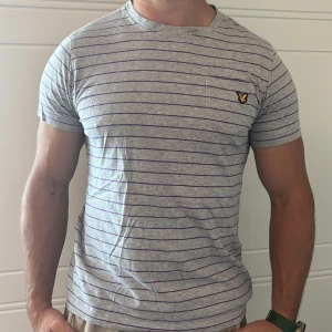 Grå randig t-shirt från Lyle & Scott - Snygg grå t-shirt från Lyle & Scott med tunna lila ränder och klassisk logga på bröstet. Modellen är kortärmad och har en normal passform. Liten i storlek motsvarar en tight M. 