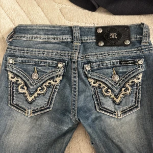 Miss Me jeans - Säljer ett par ljusblå jeans från Miss Me med snygga broderade bakfickor, blingiga knappar och slitna detaljer. Jeansen är extra low waist och i storlek 25 i midja. Midjan rakt över är 37,5cm och innerbenslängden är 81cm💓 har ett hål i indre låret och lite slitna längst ner! Köpte för 1800kr