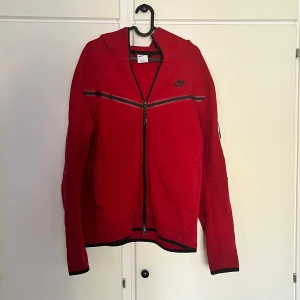 Röd Nike tech - Nike hoodie i rött med svarta detaljer och dragkedja hela vägen. Snygg sportig look med huva, svarta ränder över bröstet och diskret Nike-logga. Perfekt för dig som gillar streetstyle och vill ha något bekvämt och stilrent.