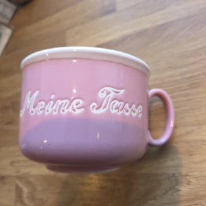 Mugg "Meine Tasse"  - Bilderna visar en rosa mugg med texten 'Meine Tasse' i vit relief. 10 cm i diameter. Muggen har en mjuk, rund form och en vit kant upptill. Perfekt för dig som vill ha en personlig och färgglad kopp till din dryck. Passar både hemma och på kontoret för en ungdomlig och trendig känsla. Lite sliten på insidan, men inget som stör användningen. 