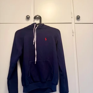 Mörkblå hoodie från Polo Ralph Lauren - Snygg mörkblå hoodie från Polo Ralph Lauren i storlek XS. Den är nästan som ny förutom att snörerna har blivit lite rosa, köpt för 1200kr tryck gärna på ”köp nu”❣️