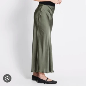 Lång grön kjol  - Snygg olivgrön långkjol i satin med lätt glansig finish och mjukt fall. Kjolen har en enkel, stilren design som sitter snyggt i midjan och faller rakt ner. Perfekt för dig som gillar en clean och trendig look. Hade beställt kjolen och det passade inte mig och glömde göra ett Retur.