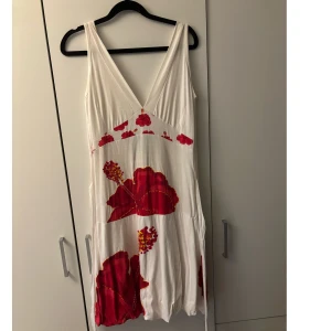 Vintage Desigual klänning❤️ - Snygg vit kortklänning från Desigual med stora röda blommor och broderad logga i ryggen. Djup v-ringning fram och bak, knytband i midjan och ärmlös design. Perfekt för sommarensdagar och ger en färgstark look🥰