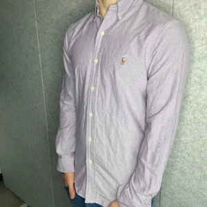 Lila skjorta från Polo Ralph Lauren - Snygg lila skjorta från Polo Ralph Lauren i slim fit-modell. Skjortan har klassisk button down-krage, broderad logga på bröstet och vita knappar framtill. Tillverkad i bomull och har långa ärmar. Perfekt för dig som gillar stilrena och tidlösa plagg.