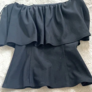 Svart offshoulder topp från Zara - Supersnygg svart offshoulder topp från Zara med volang upptill. Sitter tajt över kroppen och har en stilren, elegant look. Perfekt för dig som vill sticka ut med en trendig och chic vibe. Toppens material känns mjukt och följsamt.