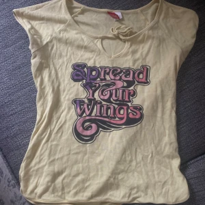 Söker Gul topp med tryck och knyt - Söker Snygg ljusgul topp från hm med coolt retrotryck 'Spread Your Wings' i lila och rosa. Toppen har korta ärmar, knyt vid halsen och en liten öppning framtill. 