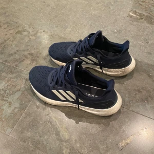 Adidas Ultraboost marinblå sneakers - Snygga Adidas Ultraboost sneakers i marinblått med klassiska vita ränder på sidorna. Ovandelen är i ventilerande syntetmaterial och sulan är vit och riktigt bekväm. Perfekta för dig som gillar sportig stil och vill ha sköna skor till vardagen. Endast en lite skada där fram till höger. 