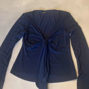 Mörkblå omlottopp från Gina Tricot - Mörkblå långärmad topp från Gina Tricot med snygg omlottdesign och knyt framtill. Djup v-ringning och mjukt, stretchigt material som sitter skönt på kroppen. Perfekt för dig som gillar stilrena och trendiga plagg med en twist.