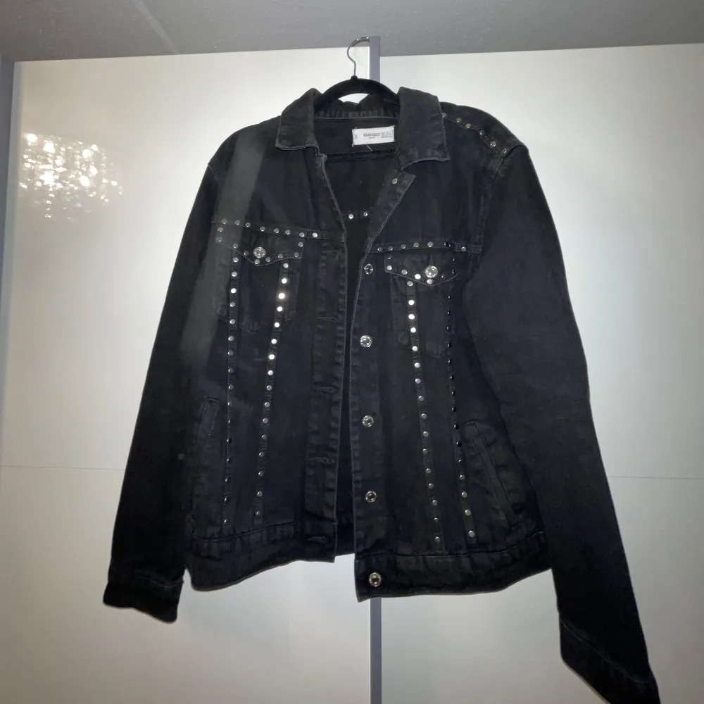 Snygg svart jeansjacka från mango med coola nitar längs framsidan och axlarna. Jackan har en klassisk krage och knappar framtill. Perfekt för att ge din outfit en edgy touch.. Takit.