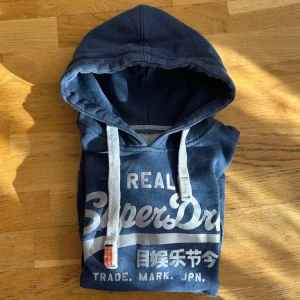 Blå hoodie från Superdry - Säljer en snygg blå hoodie från Superdry med vit/silvrig  logga på framsidan. hoodien är lite chunky vilket många gillar om du kollar på snörerna så ser man det just den här färgen och designen är väldigt svår att hitta och säljs inte längre på hemsidan jag skulle säga att hoodien passar någon som är cirka 170cm