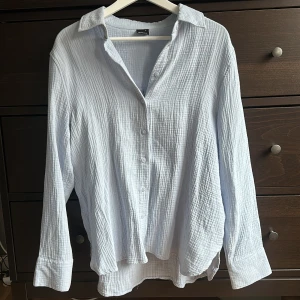 Ljusblå skjorta  - Säljer en fin ljusblå skjorta i ett lätt och luftigt material. Skjortan har långa ärmar och knappar framtill. Endast använd någon enstaka gång så den är i nyskick. 