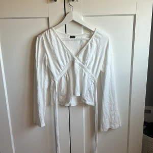 Vit omlottopp från Gina Tricot  - Säljer en vit omlottopp från Gina Tricot i storlek XS. Toppen har långa ärmar och ett knytband i midjan för en justerbar passform. Använd ett fåtal gånger så tröjan är i väldigt bra skick!!