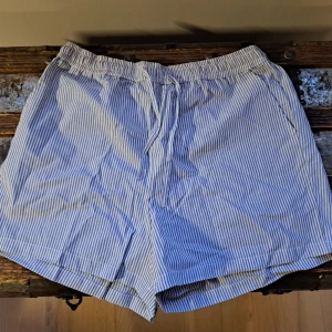 Randiga blå och vita shorts - Säljer ett par bekväma blå och vitrandiga shorts med elastisk midja och dragsko. Perfekta för en avslappnad dag i solen. De har en klassisk look med två sidofickor.