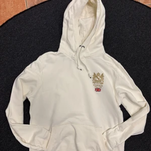 Vit hoodie från Morris - Säljer en stilren vit hoodie från Morris med broderad logotyp och brittisk flagga på bröstet. Tröjan har en justerbar huva med snörning och en praktisk magficka. Perfekt för en avslappnad stil.