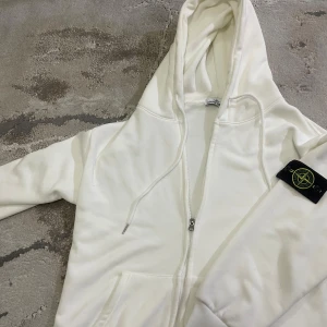Vit hoodie från Stone Island - Säljer en stilren vit hoodie från Stone Island med klassisk logga på ärmen. Tröjan har en dragkedja framtill och en justerbar huva. Perfekt för en avslappnad look och har en väldigt skön pels på insidan av Hoodien.