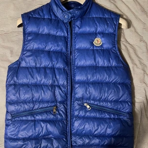 Blå dunväst från Moncler - Snygg blå dunväst från Moncler med dragkedja och två fickor framtill. Perfekt för kyligare dagar och ger en stilren look. Moncler-loggan pryder bröstet för en exklusiv touch. Storlek 2 S/M. Pris kan diskuteras. 