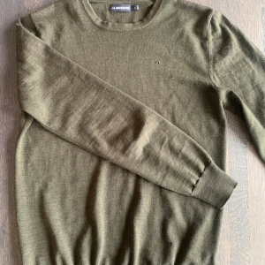 Olivgrön merinoulltröja från J.Lindeberg - Säljer en stilren olivgrön tröja från J.Lindeberg i mjuk merinoull. Tröjan har en rund halsringning och långa ärmar, perfekt för kyligare dagar. Diskret logga på bröstet ger en elegant touch. Passar både till vardags och mer uppklädda tillfällen.