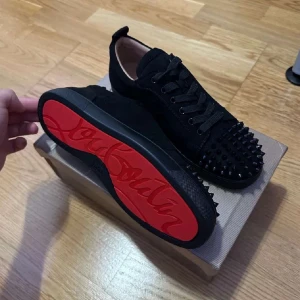 Svarta loubs med nitar och röd sula - Snygga svarta sneakers från Christian Louboutin med ikonisk röd sula och coola nitar på tån. Skorna har snörning och är tillverkade i mocka för en lyxig känsla. Priset kan diskuteras.