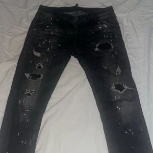 Svarta slitna jeans från Dsquared2 - Säljer ett par svarta jeans från Dsquared2 med coola slitningar och färgstänk. Jeansen har en normal passform och är perfekta för en edgy look. Märkesdetaljer syns på baksidan.