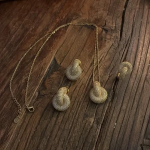 Guldigt smyckeset från Edblad  - Elegant smyckeset i guld från Edblad med ett halsband och matchande örhängen och ring.Båda delarna har en unik, vriden design med glittriga detaljer som ger en lyxig känsla. Perfekt för att lyfta vilken outfit som helst.