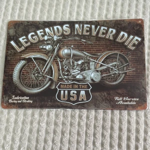 Metallskylt med motorcykelmotiv - Cool metallskylt med texten 'Legends Never Die' och en klassisk motorcykel i fokus. Skylten har en vintage-look med texten 'Made in the USA' och detaljer om smörjning och service. Perfekt för garage eller man cave!