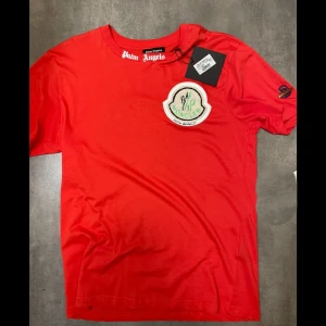 Palm Angels t shirt - Snygg röd t-shirt från Palm Angels x moncler  med ett stort tryck på ryggen och en broderad patch på bröstet. T-shirten har korta ärmar och en rund halsringning. Perfekt för dig som vill sticka ut med stil!