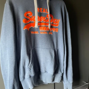 Blå hoodie från SuperDry - Säljer en snygg blå hoodie från SuperDry med en stor orange logga på bröstet. Tröjan har en klassisk känguruficka och vita dragsnören i huvan. Perfekt för en avslappnad stil.