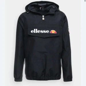 Svart vindjacka från Ellesse - Säljer en svart vindjacka från Ellesse med stor logga på bröstet och en patch på ärmen. Jackan har en dragkedja vid halsen och en stor ficka framtill. Jacksn är använd fåtals gånger original pris 1299kr