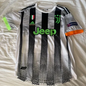 Juventus fotbollströja - Snygg Juventus fotbollströja från Adidas med svartvitt randigt mönster och gröna detaljer. Tröjan har korta ärmar med en unik design där ena ärmen är grön och den andra orange. Perfekt för fotbollsfans!