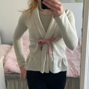 Beige kofta från Odd Molly - Säljer en söt beige kofta från Odd Molly med rosa knytband i midjan och volangdetalj nedtill. Koftan är långärmad och tillverkad i 100% bomull. Perfekt för en mysig dag!
