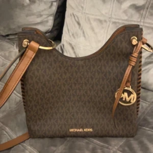 Brun axelväska från Michael Kors - Snygg brun axelväska från Michael Kors med klassiskt MK-mönster. Väskan har justerbar axelrem och dekorativt hängsmycke. Insidan är rymlig med flera fack och stängs med magnetknapp. Perfekt för att bära med sig det viktigaste med stil.
