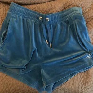 Blå velourshorts från Juicy Couture - Snygga blå velourshorts från Juicy Couture med glittrande stenar som bildar texten 'Juicy' på baksidan. Shortsen har en elastisk midja med dragsko och praktiska fickor med dragkedja. Perfekta för en avslappnad och stilren look.