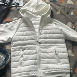 Vit quiltad jacka med huva - Säljer en stilren vit quiltad jacka med huva. Jackan har dragkedja framtill och två praktiska fickor med dragkedjor. Perfekt för kyligare dagar.