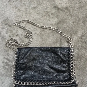 Svart axelväska från Tiamo - Snygg svart axelväska från Tiamo i skinn med en kedja som axelrem. Väskan har en stilren design med dragkedja och kedjedetaljer runt kanterna. Perfekt för att ge en edgy touch till din outfit.